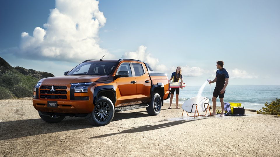 L200 laranja estacionada numa praia, com duas pessoas a lavar pranchas de surf