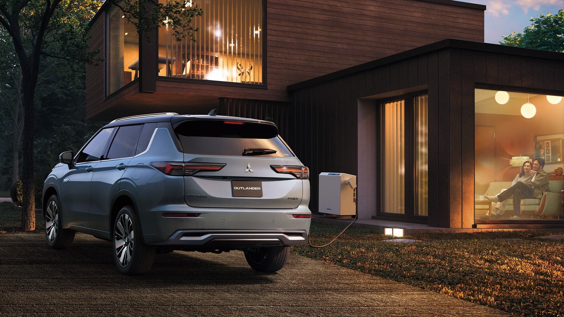 Mitsubishi Outlander PHEV prateado ligado a um carregador bidirecional, à entrada de uma casa ao anoitecer
